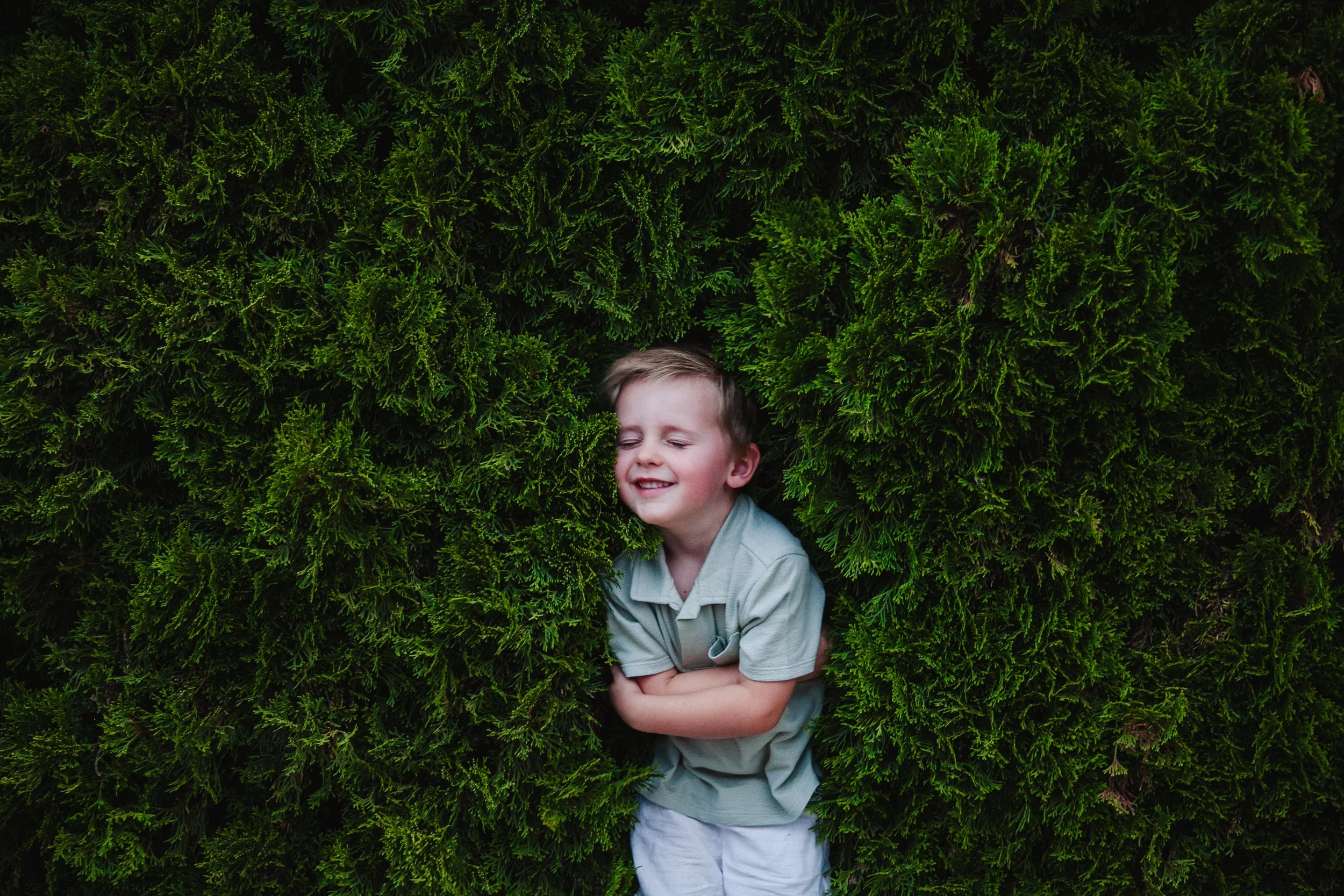 A young boy with light-colored hair and a light gray shirt is smiling with his eyes closed as he stands among green, lush hedge bushes, holding his crossed arms in front of him.
