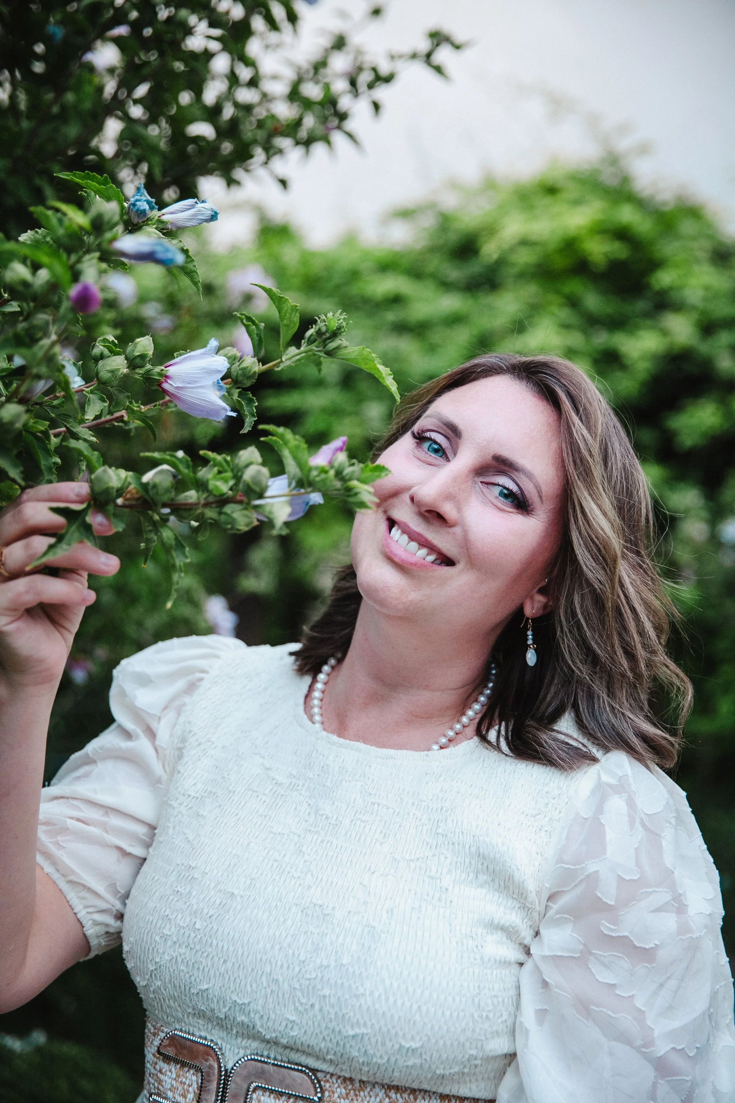 A woman with blue eyes and brown hair smiling in a garden, holding a branch with purple and white flowers.