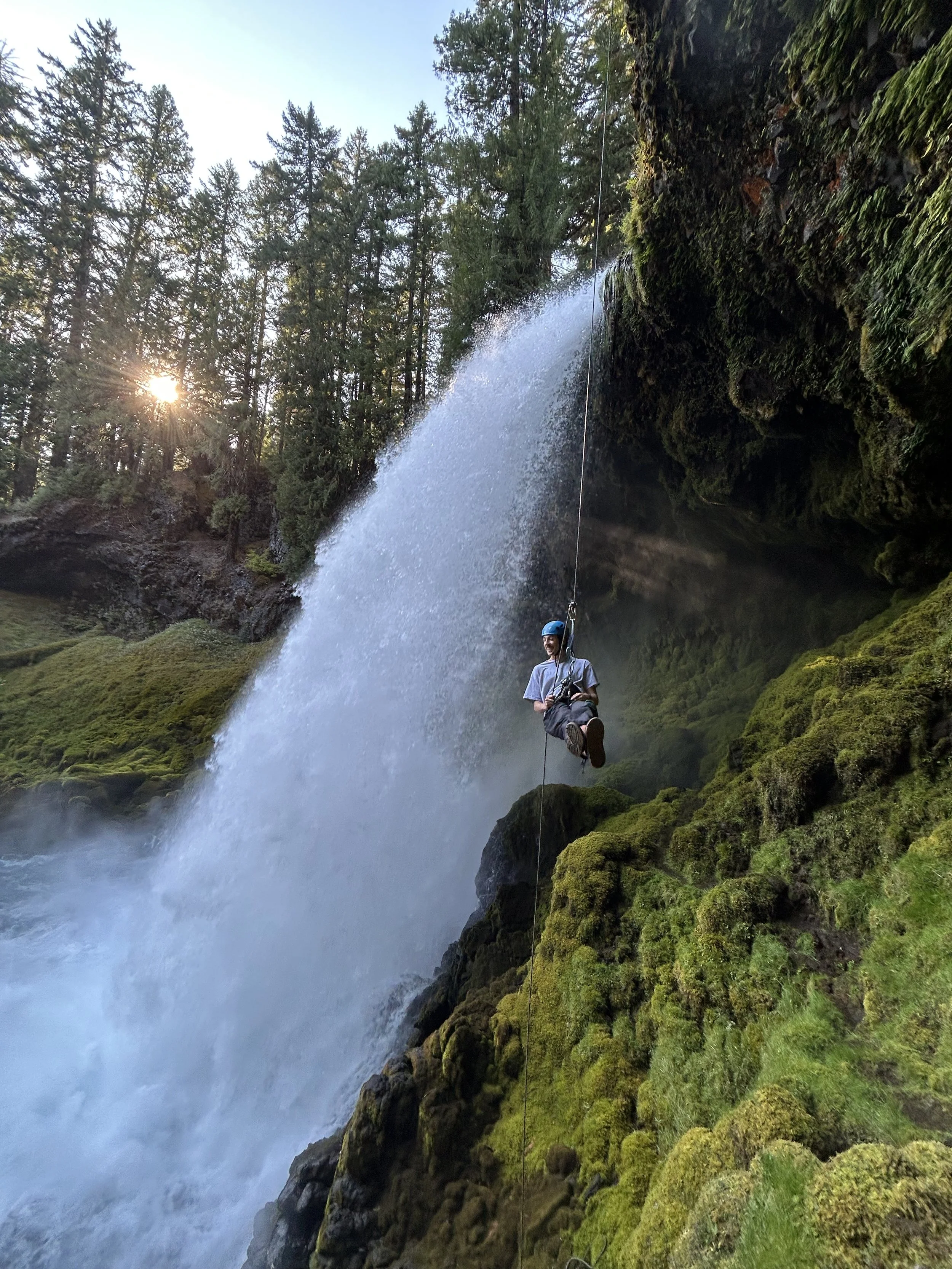 A man rappelling, stopped halfway down tand looking out at a waterfall in the forest.