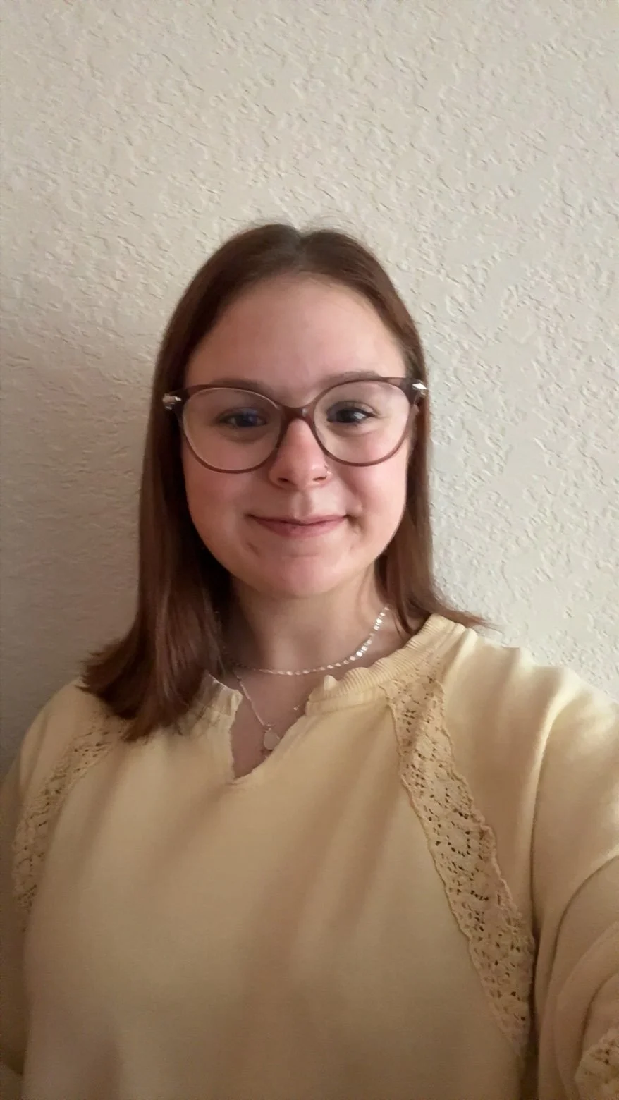 A young woman sitting on a park bench wearing a black hat and a red plaid scarf, with green trees and a few buildings in the background.