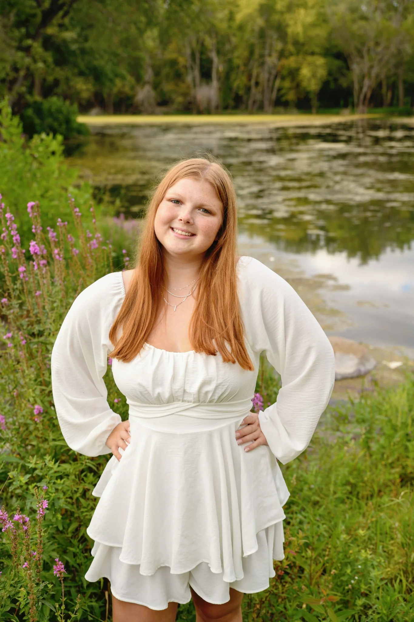 A young woman with long red hair, smiling and posing in front of an apple tree with green leaves and red apples.