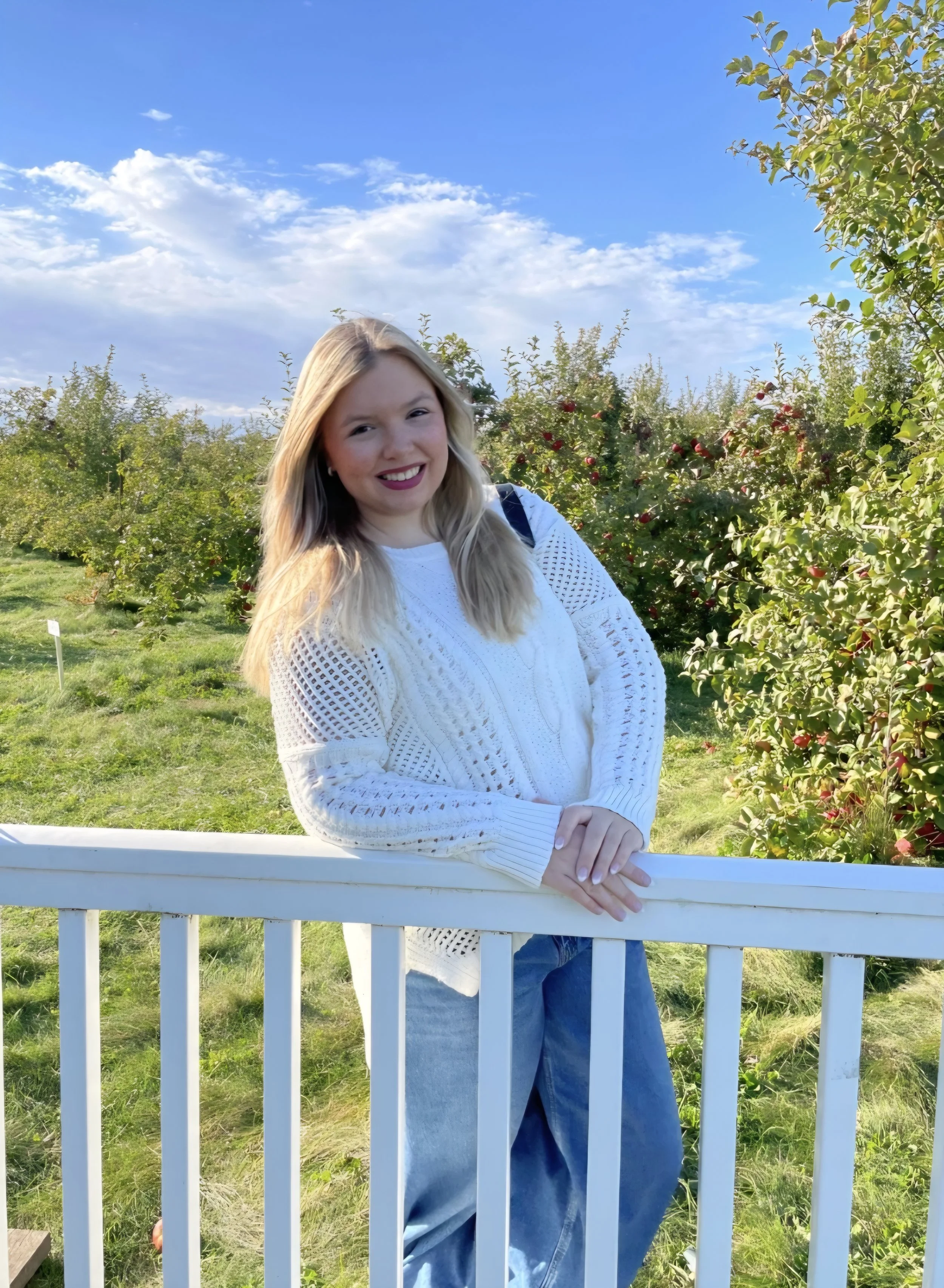 A smiling young woman in a white hoodie and blue jeans standing on a boat with a mountainous landscape and a turquoise lake in the background.