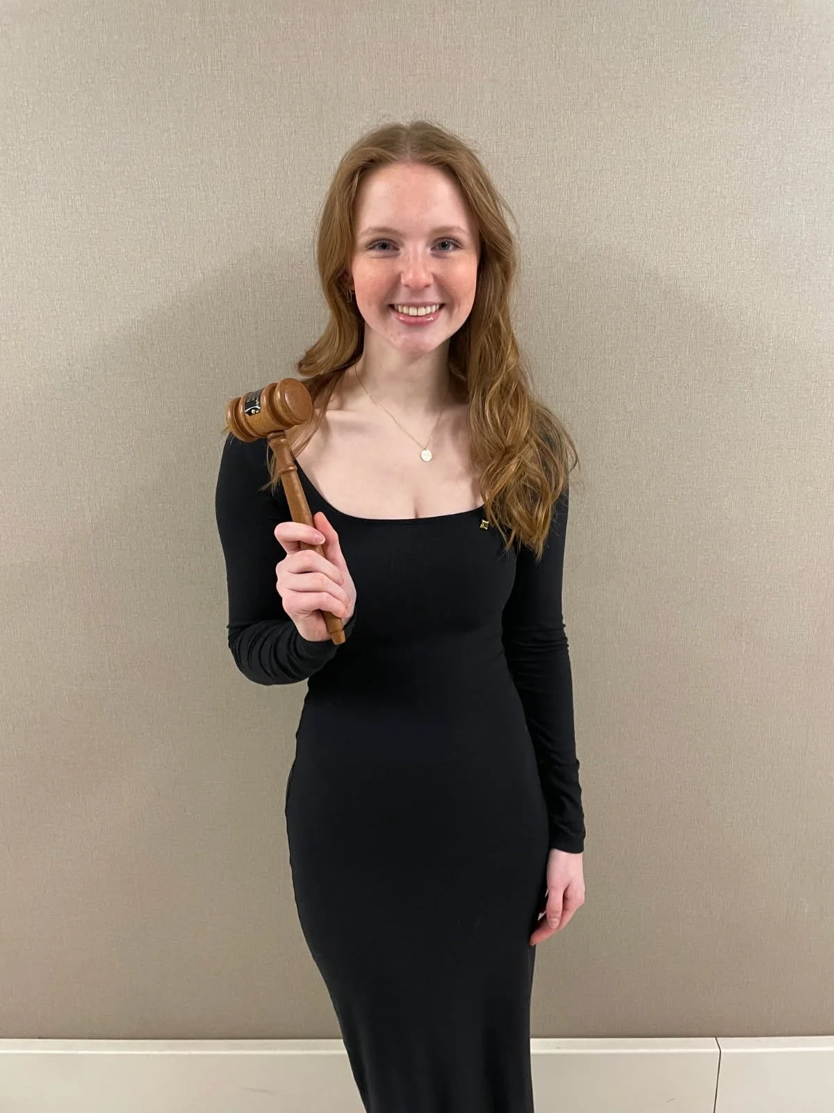 A young woman with brown, wavy hair smiles outdoors while holding a judge's gavel. She wears a white dress with purple floral patterns and small earrings. Background includes green trees and sunlight.