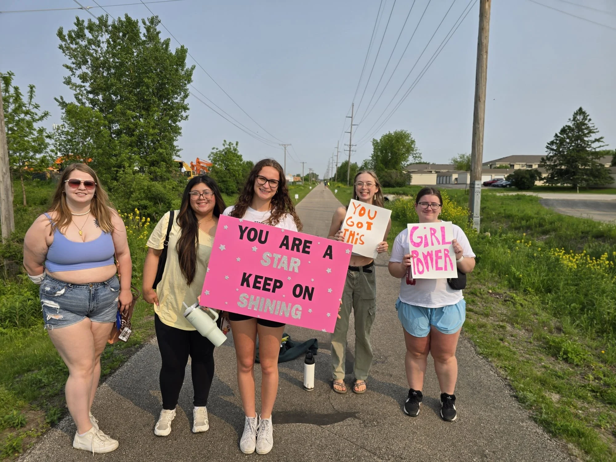 Two women holding a bright green poster with the words 'GOGO GOGO' and playful images of hats, stars, and megaphone, outdoors in snowy weather, with trees and other people in the background.