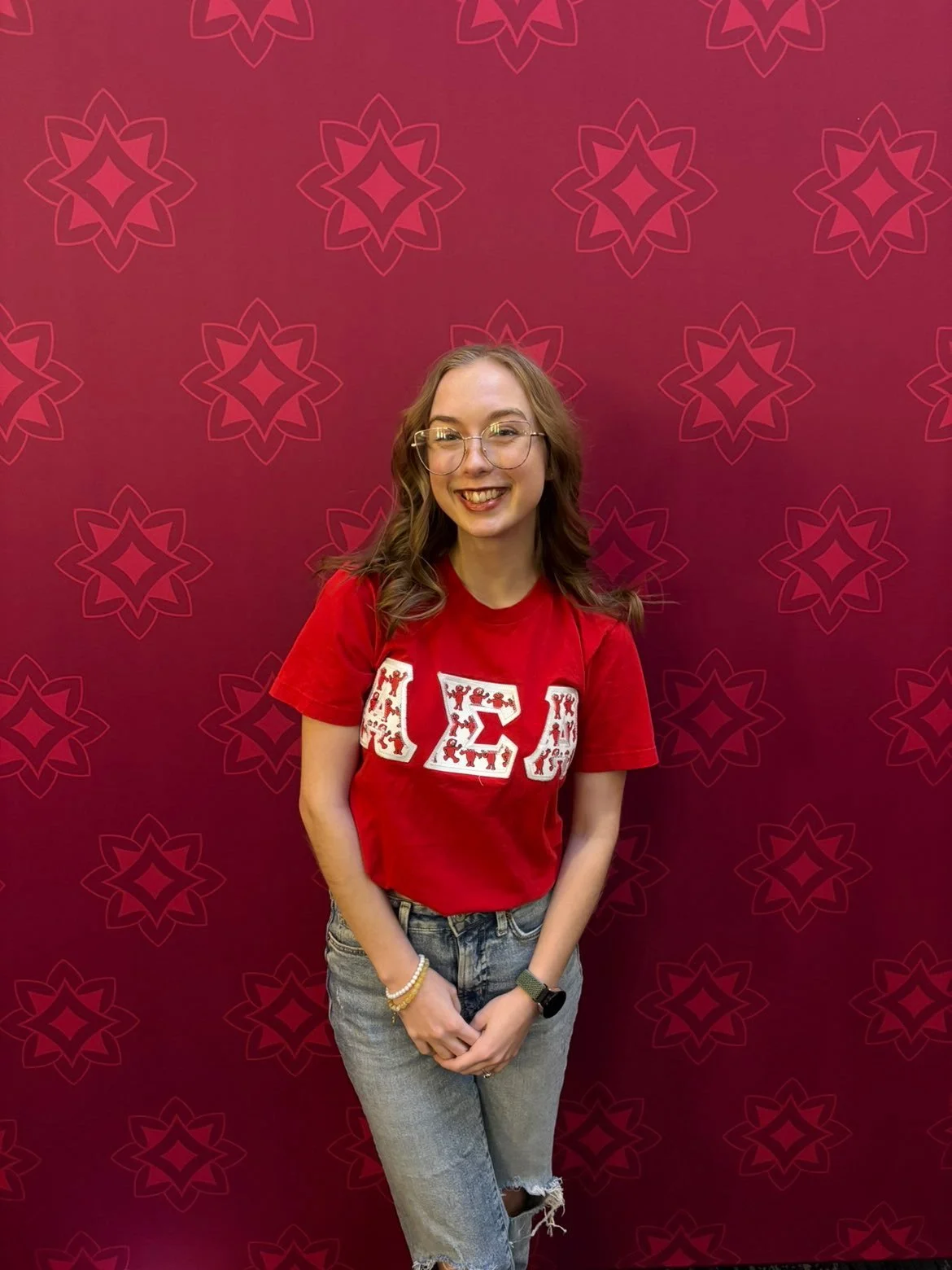 A young woman with glasses and shoulder-length hair, wearing a white textured dress and white high-heeled sandals, stands smiling in front of a beige wall with a colorful abstract painting partially visible on the left.