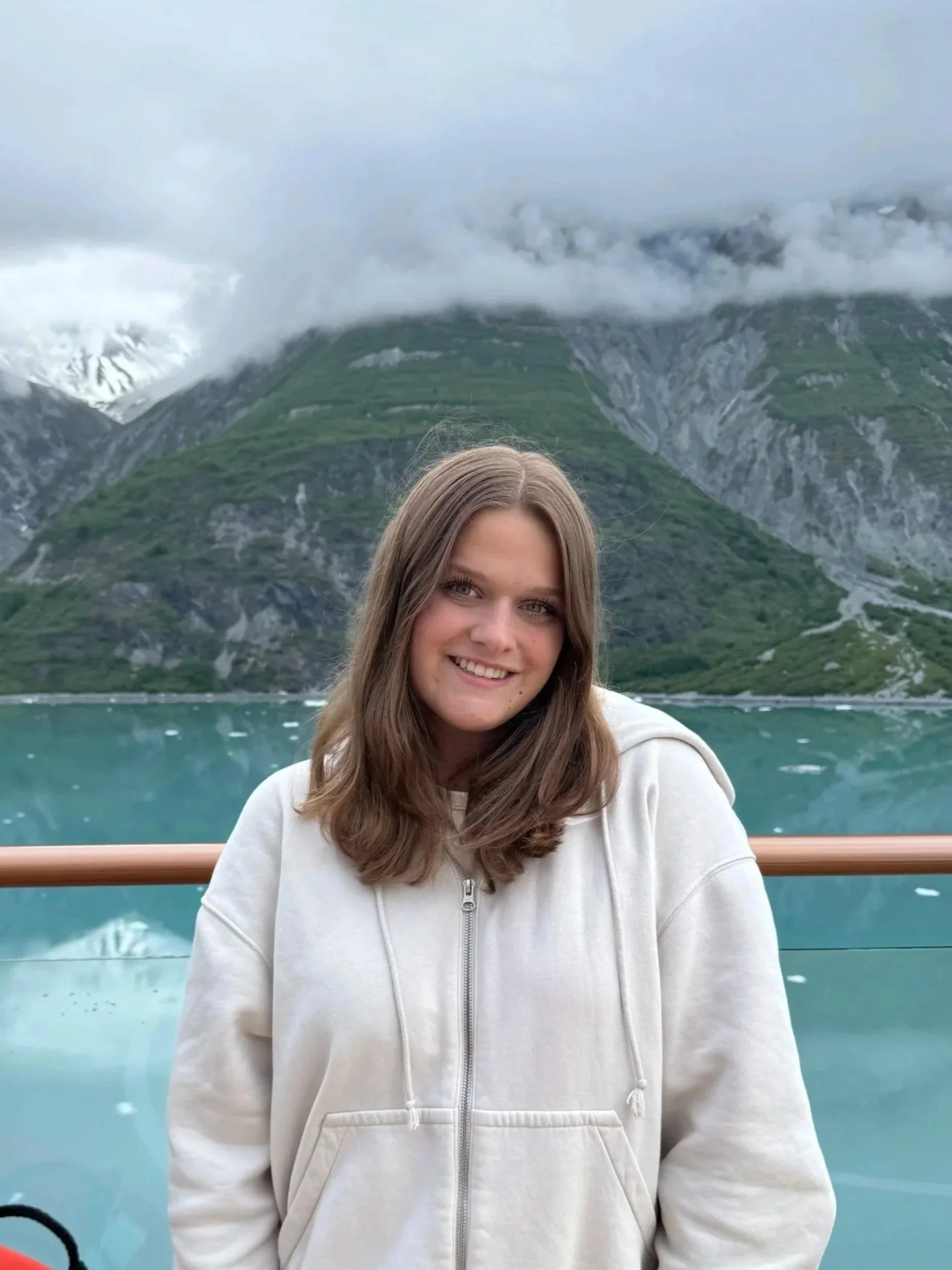 A woman with shoulder-length blonde hair smiling, wearing a maroon long-sleeve top and black pants, standing on a city bridge with city skyscrapers in the background.