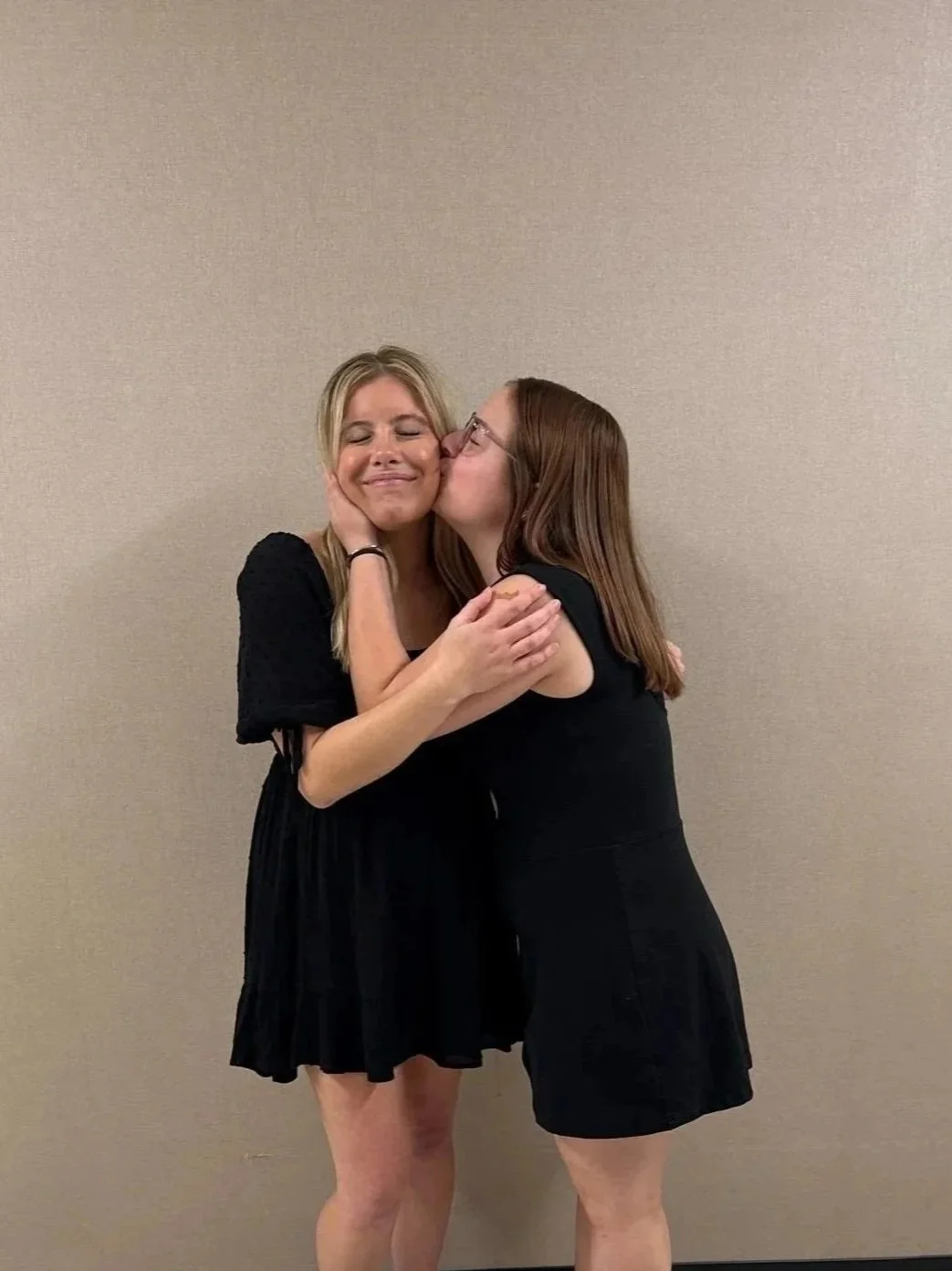 Two young women wearing white dresses hugging and smiling against a beige wall.