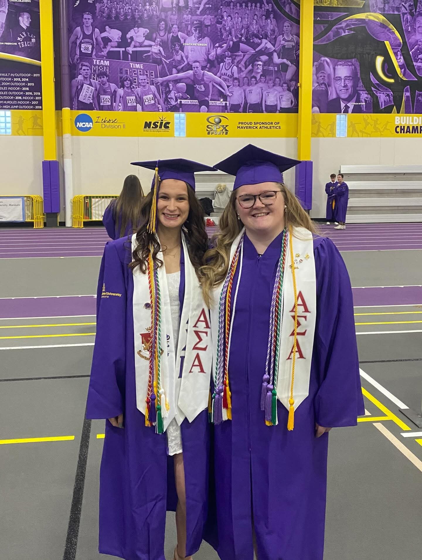 Two women sharing a hug and kiss in front of a green hedge wall with a neon sign that reads 'AFV Central'.