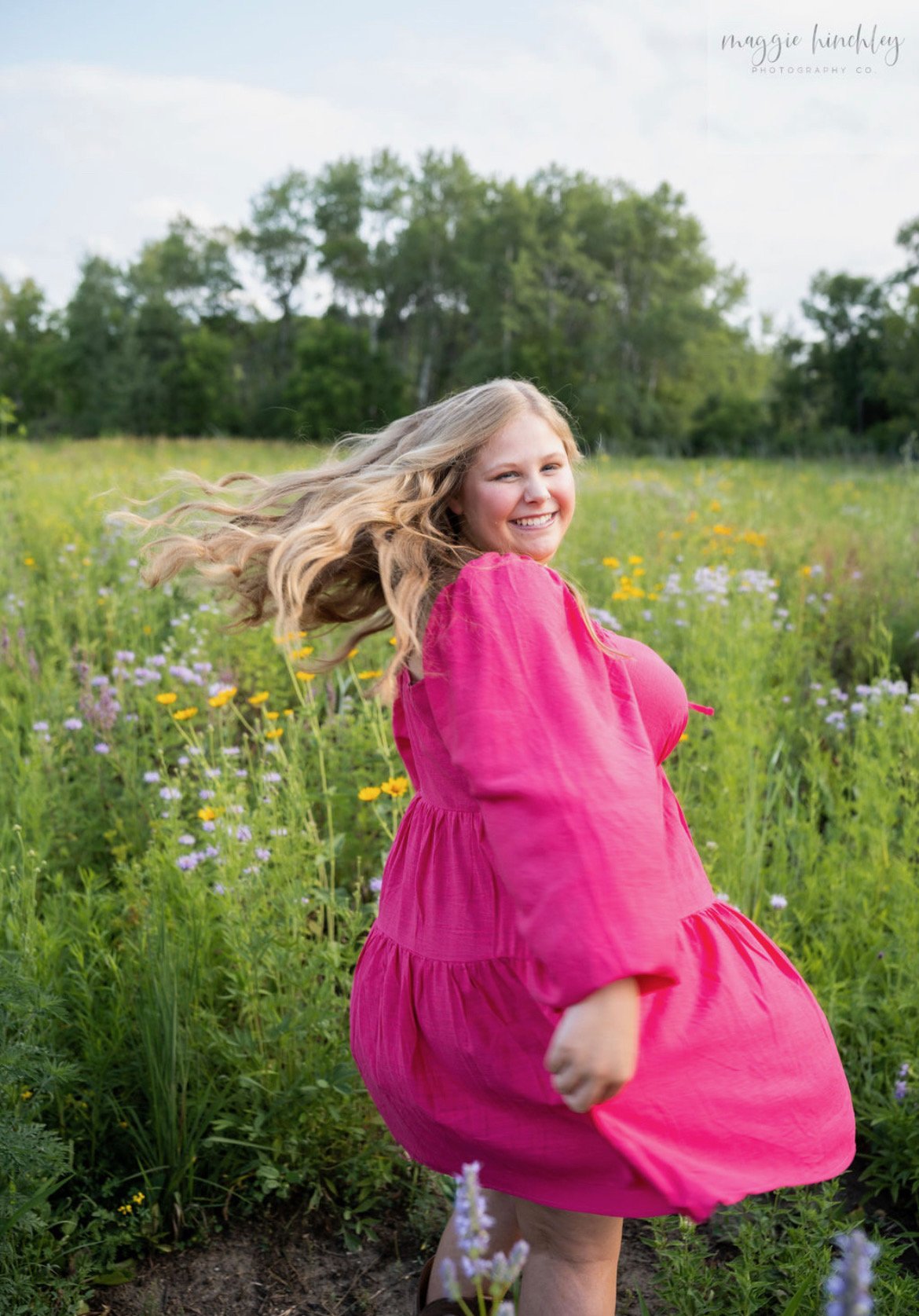 A young woman with long, wavy brown hair smiling, wearing a light-colored, sleeveless dress with polka dots and a small necklace, standing outdoors against a textured gray wall.