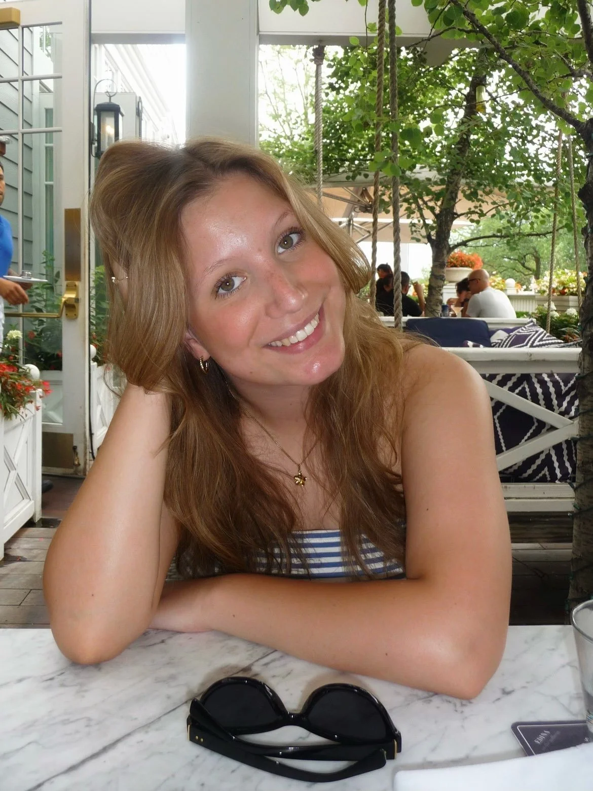 A young woman with glasses and a braid, smiling outdoors on a windy day, wearing a brown sweatshirt with "SENIOR 23" and a black crossbody bag.