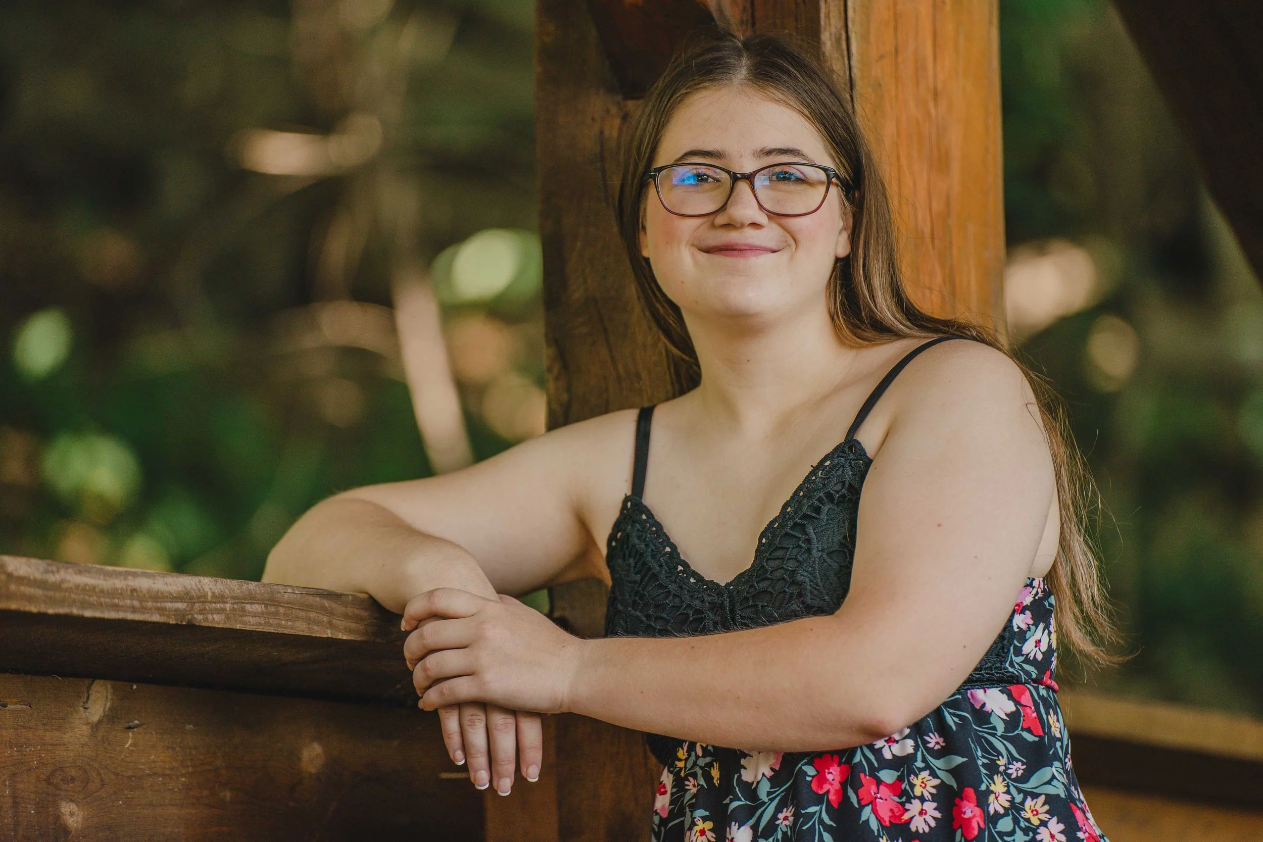 A young woman with glasses and a braid, smiling outdoors on a windy day with a rocky hillside and blue sky in the background, wearing a brown sweatshirt that says 'SENIOR 23' and carrying a black bag.