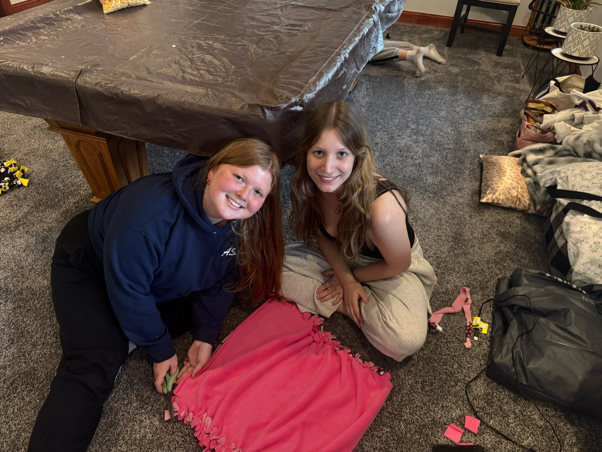 Group of young women sitting on the floor around colorful pet beds with paw prints and blankets in a room with gray carpet and chairs.