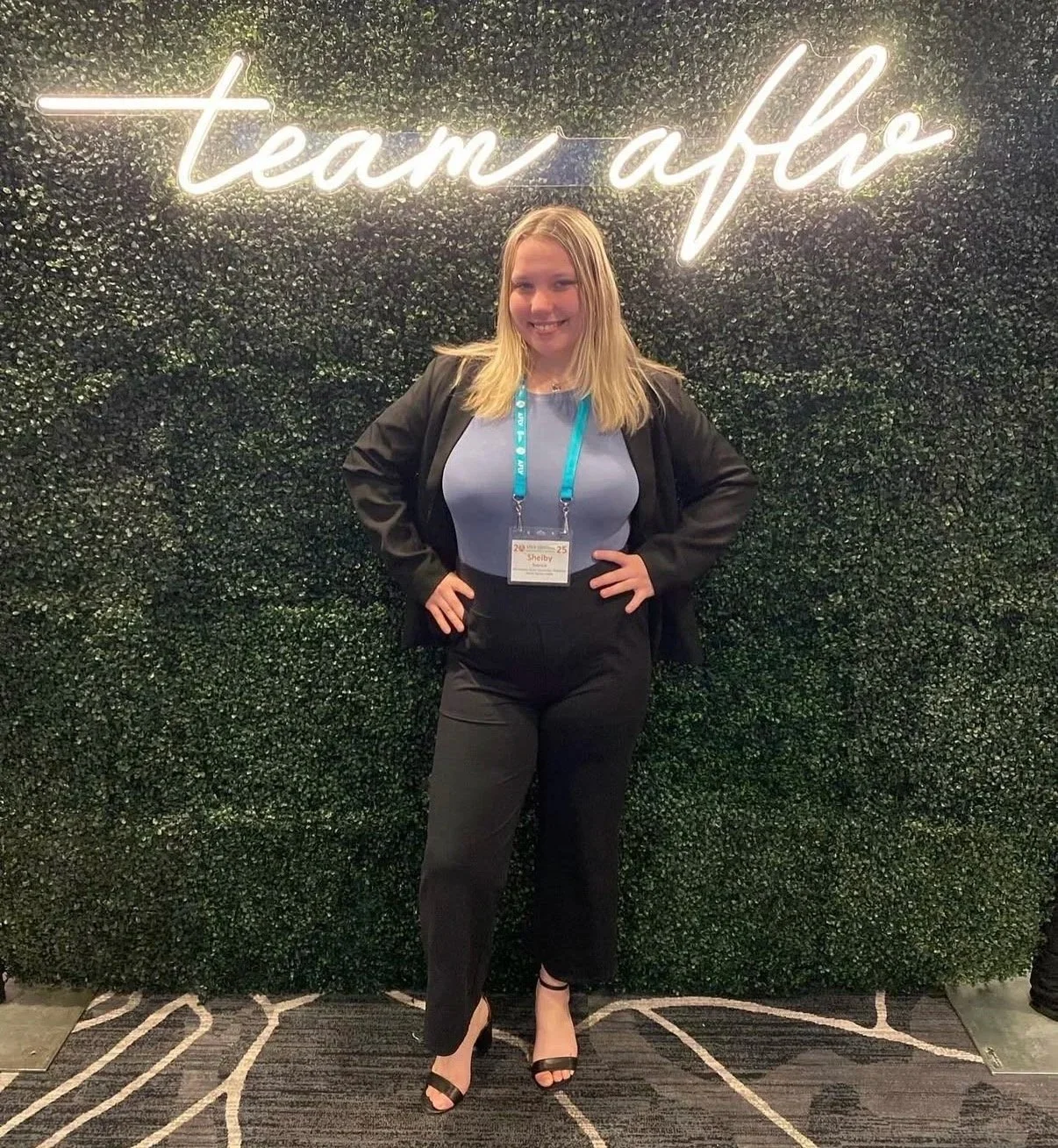 A young woman with long brown hair, smiling, standing in front of a large indoor sports arena filled with seated spectators and a stage with large red screens and purple hanging decorations.