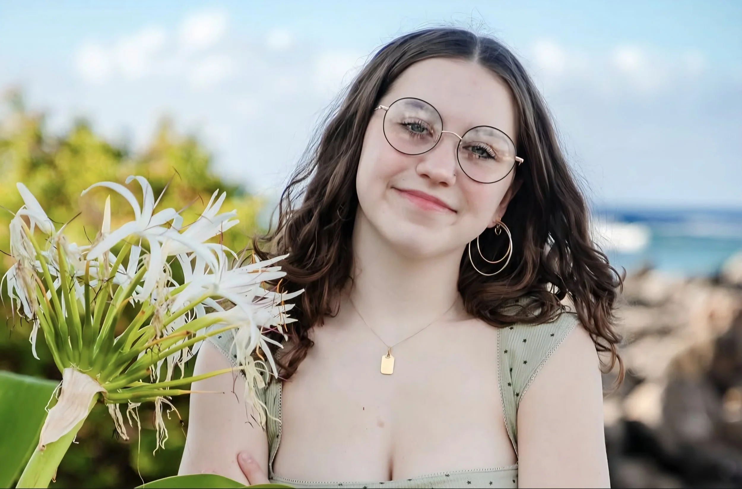 A young woman with long red hair wearing a pink dress standing in a field at sunset, smiling at the camera.