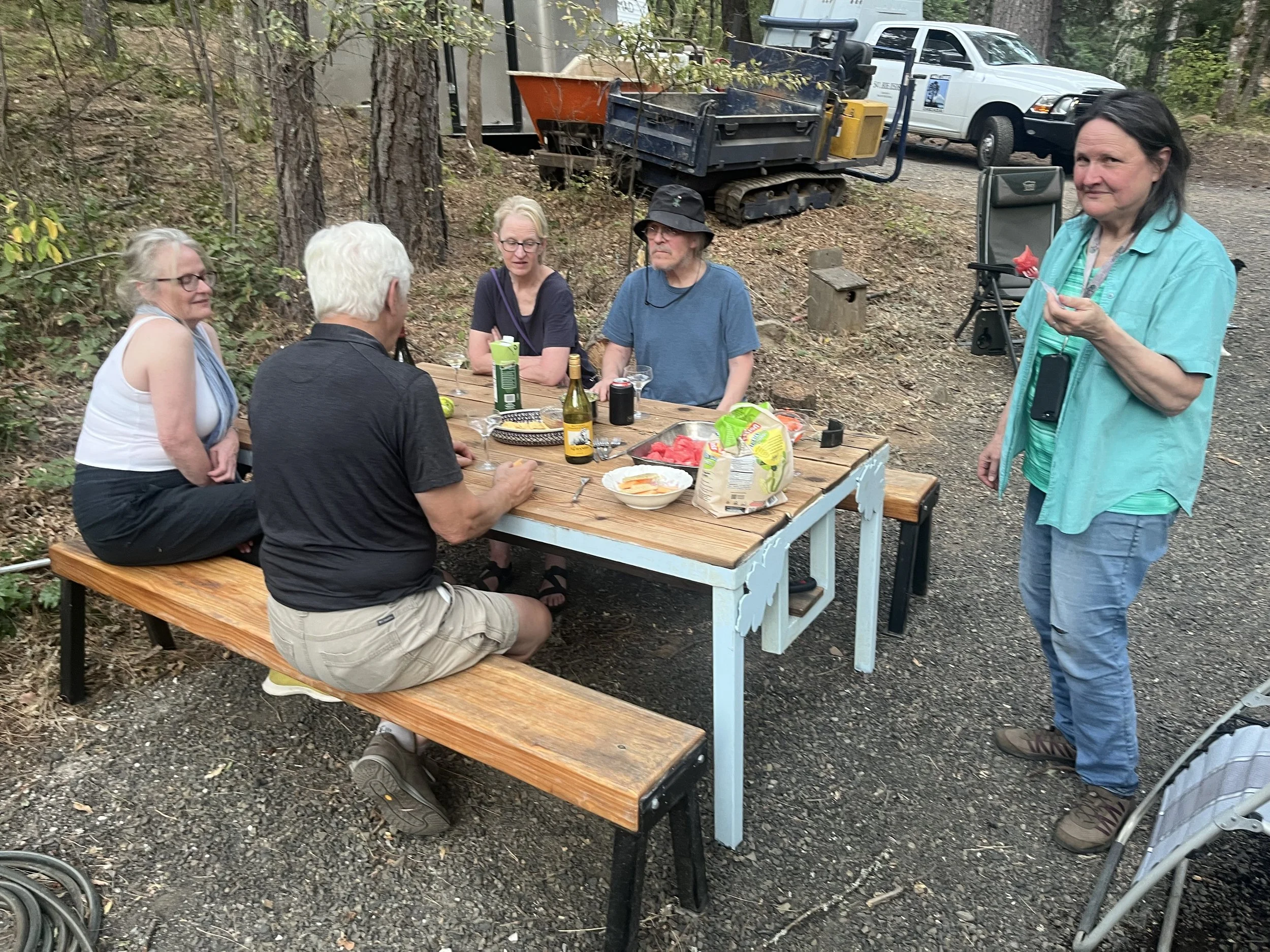 A group of five people sitting and standing around a picnic table outdoors in a wooded area, with equipment and vehicles visible in the background.