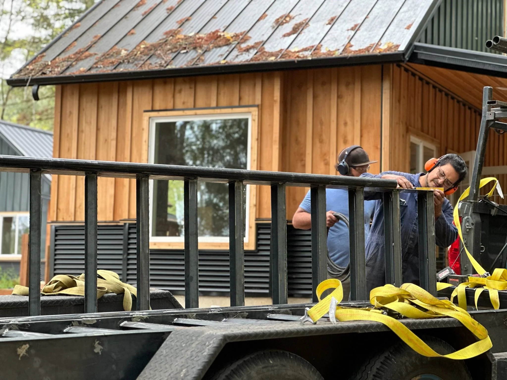 Construction workers working on a house with wooden siding, metal roof, and large window, using safety harnesses and equipment.