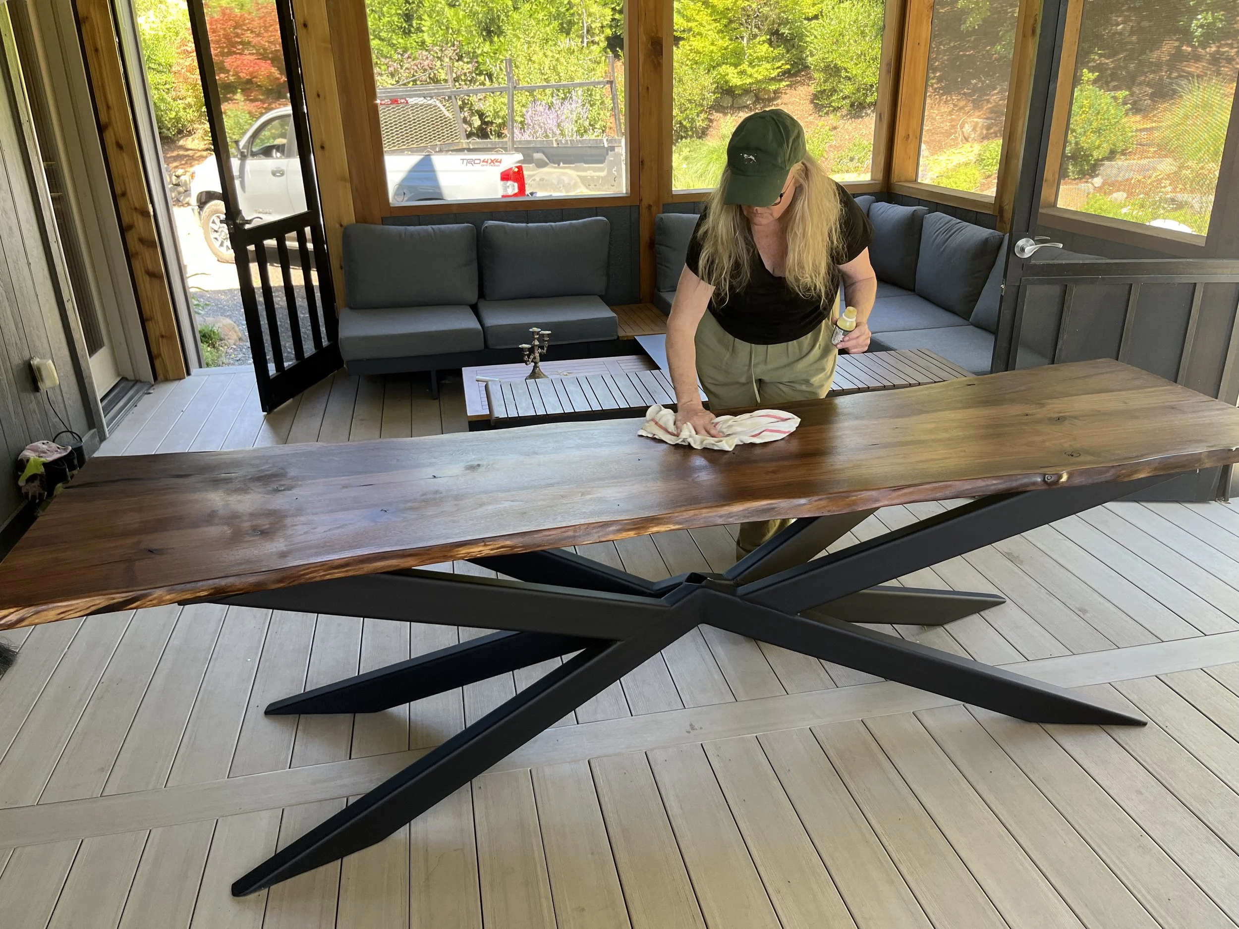 A woman cleaning a wooden table on a screened porch with outdoor views of trees, a white truck, and a patio area.