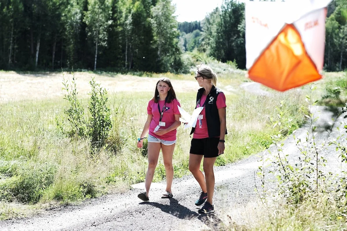 To kvinner går tur på en grusvei i et grønt landskap med trær i bakgrunnen, den ene holder en kart. En orange og hvit telt eller markeringsbøyle er delvis synlig i forgrunnen til høyre.