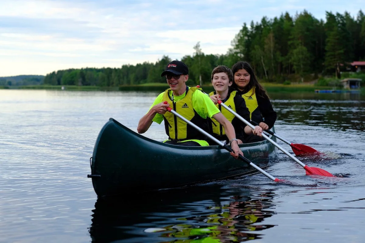 Tre personer i en kano på en rolig elv, omgitt av grønt skoglandskap, alle kledd i svart og gult, smilende og nyter utendørs kanoaktiviteter.