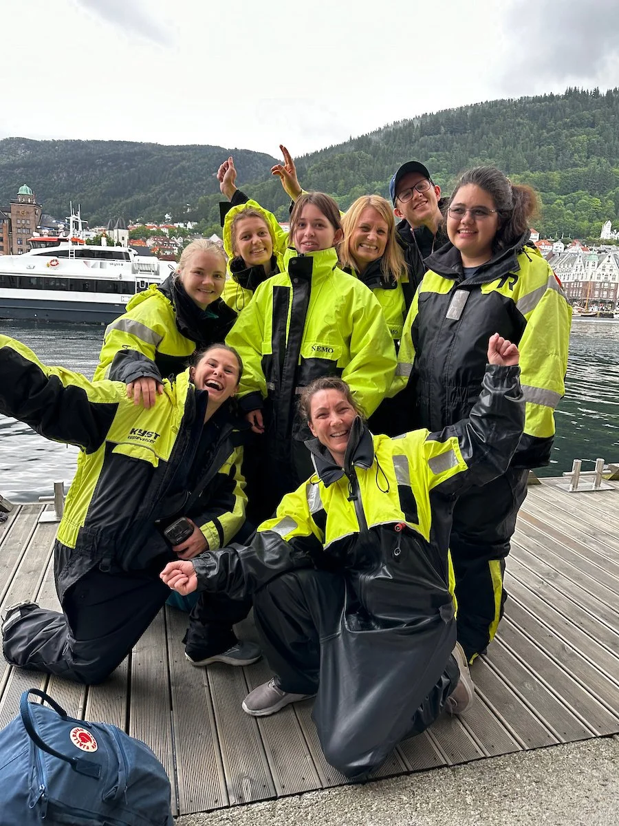 To unge kvinner ligger på gress, ser mot kamera, smiler, de har på seg lyse gule t-skjorter med sponsorlogoer. Bakgrunn med grønn vegetasjon og en blå himmel.