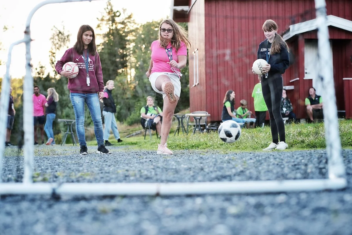 Tre jenter som spiller fotball, med to av dem holder en ball hver, mens den midterste trener eller slår ballen mellom beina på en fotballbane utendørs.