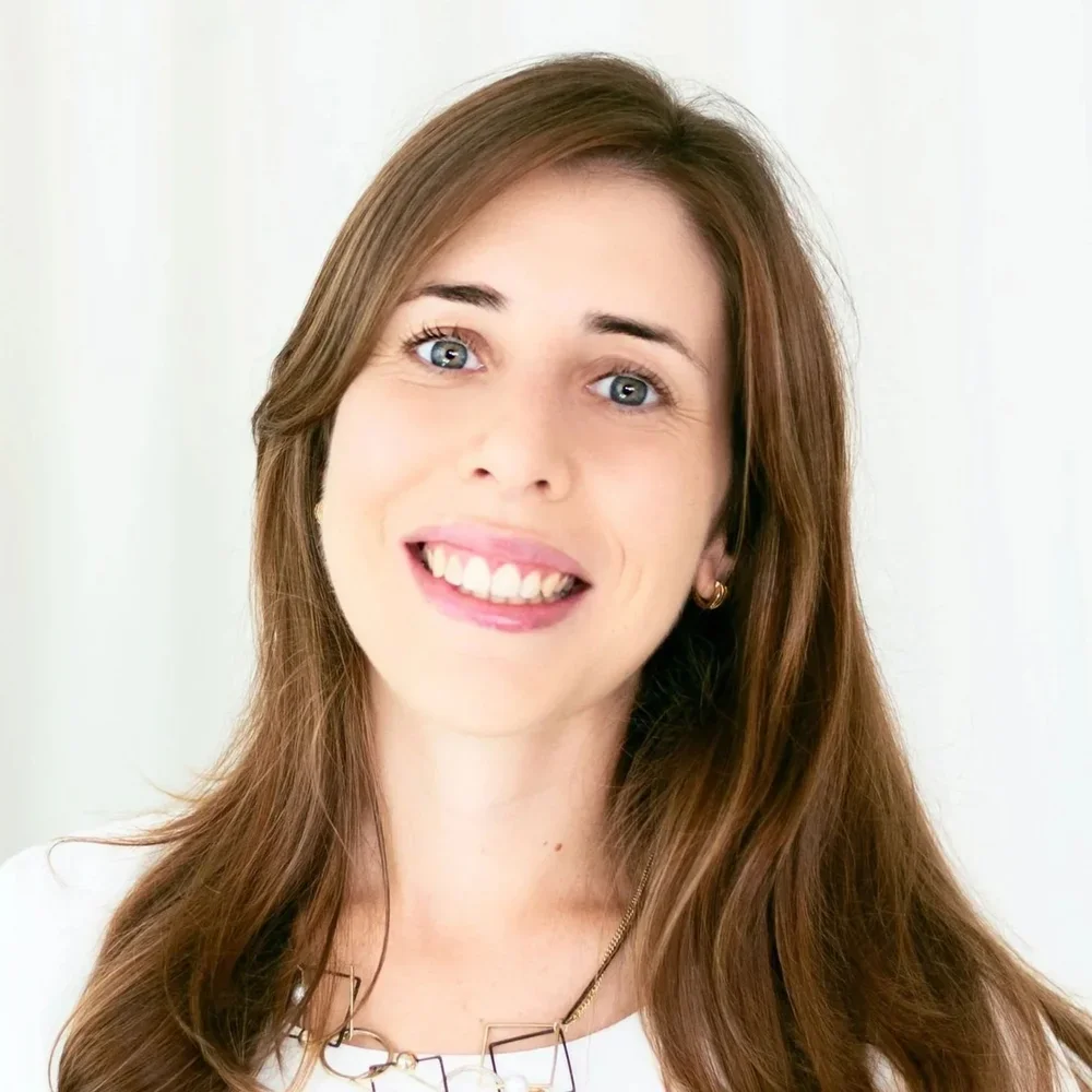 Close-up of a woman with long brown hair, blue eyes, smiling, wearing earrings and a white top, against a white background.