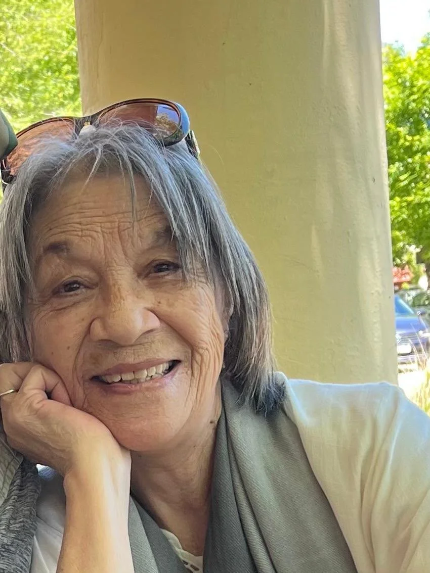 Smiling elderly woman with gray hair, wearing glasses on her head, sitting outdoors with green trees and a car in the background.