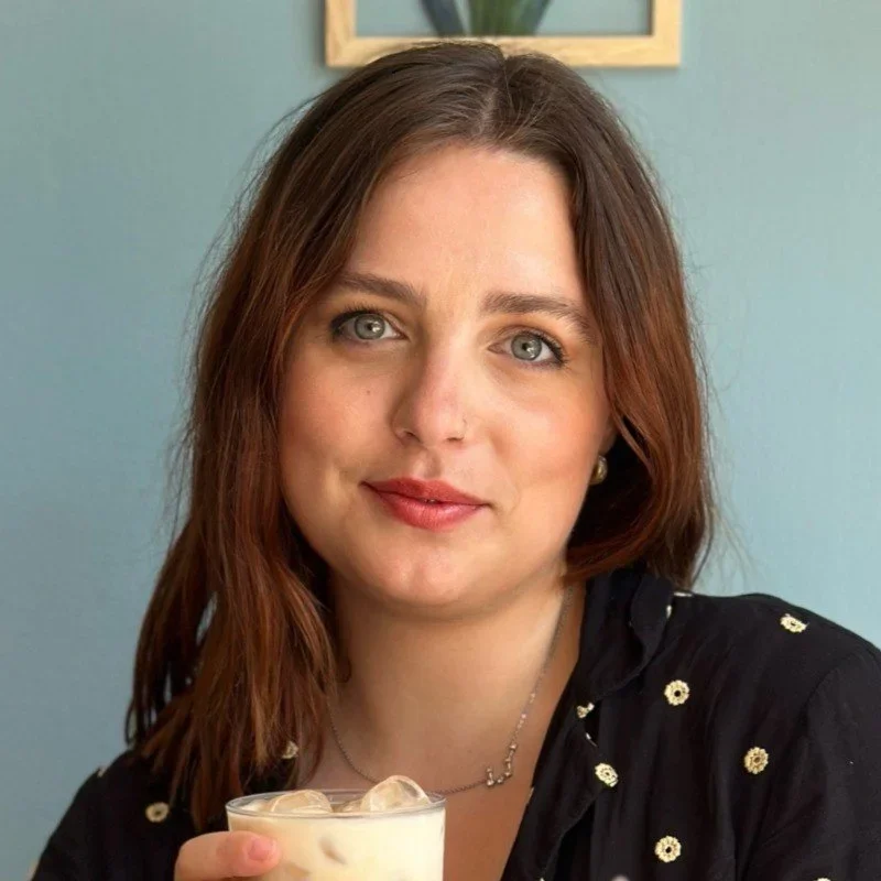 A woman with shoulder-length brown hair and fair skin, wearing a black top with small floral details and a silver necklace, holding a glass of iced coffee, seated indoors in front of a light turquoise wall with a picture frame.