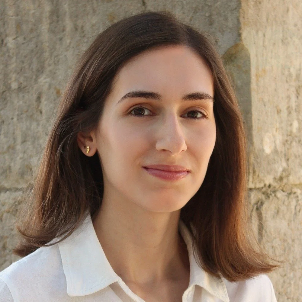 Portrait of a young woman with shoulder-length brown hair, wearing a white collared shirt, standing outdoors against a stone wall.