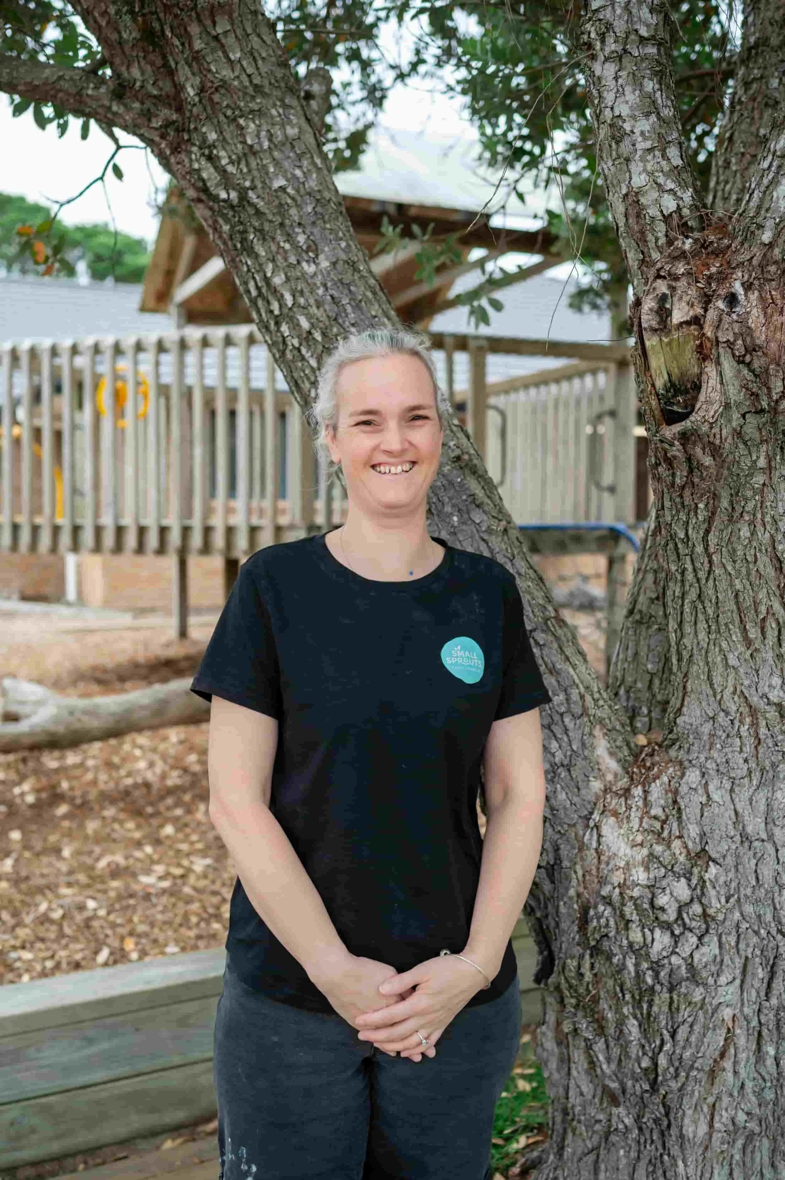 A woman with gray hair and a black T-shirt standing outdoors, smiling, with two large trees and a wooden structure in the background.
