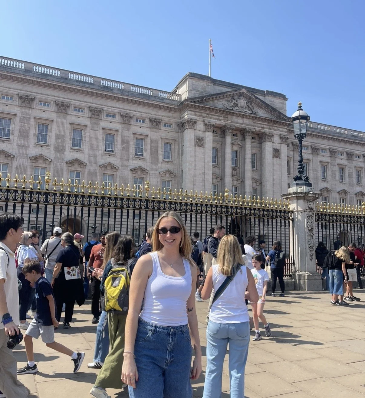 A woman with blonde hair, sunglasses, a white tank top, and blue jeans smiling in front of Buckingham Palace in London, with a crowd of tourists around her.