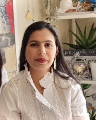 A woman with long dark hair, wearing a white blouse, standing in a cozy room with decorative shelves and plants.