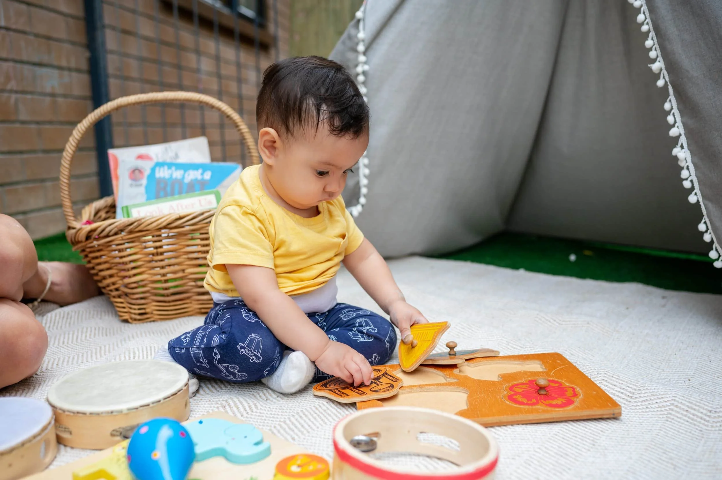 A small sprouts baby sits on a mat, playing with wooden toys near a basket of books and a tent at daycare.