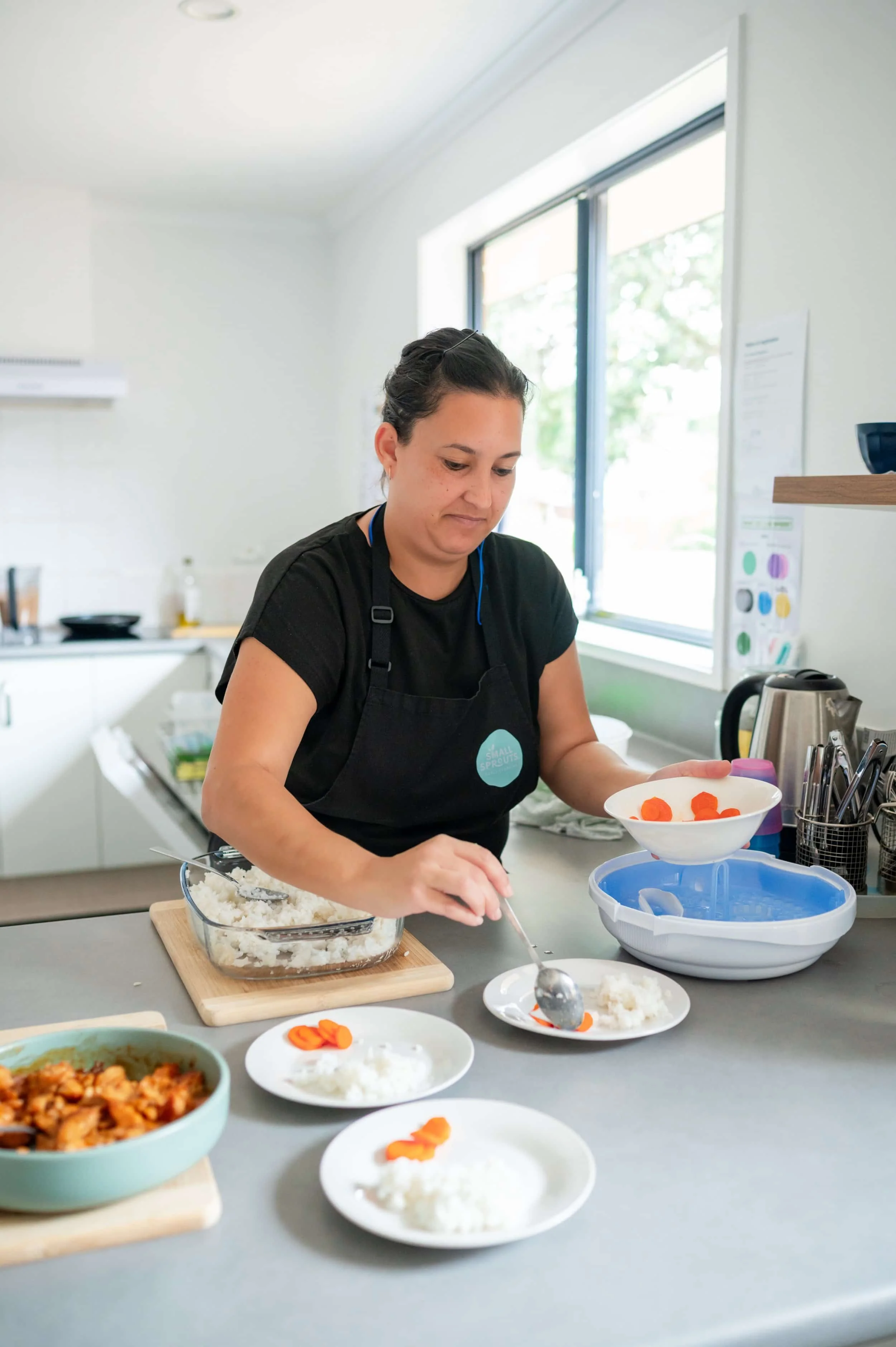 A small sprouts chef cooking, serving rice and carrots onto plates near a bright kitchen window.