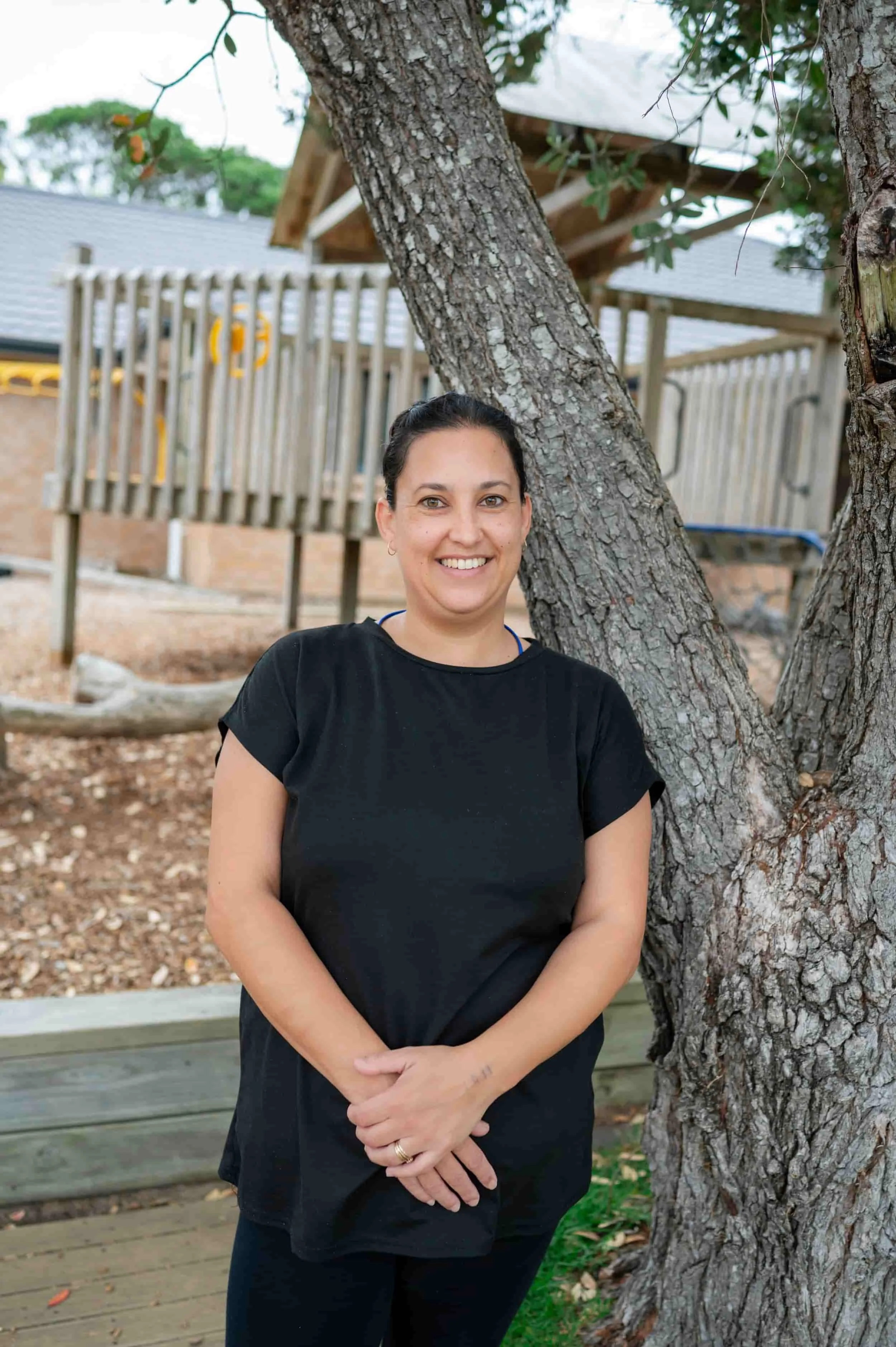 A woman with dark hair, wearing a black T-shirt, stands outdoors next to a tree with a textured trunk. She is smiling and has her arms crossed, touching her hands together at her waist. There is a wooden deck and a fenced play area in the background.