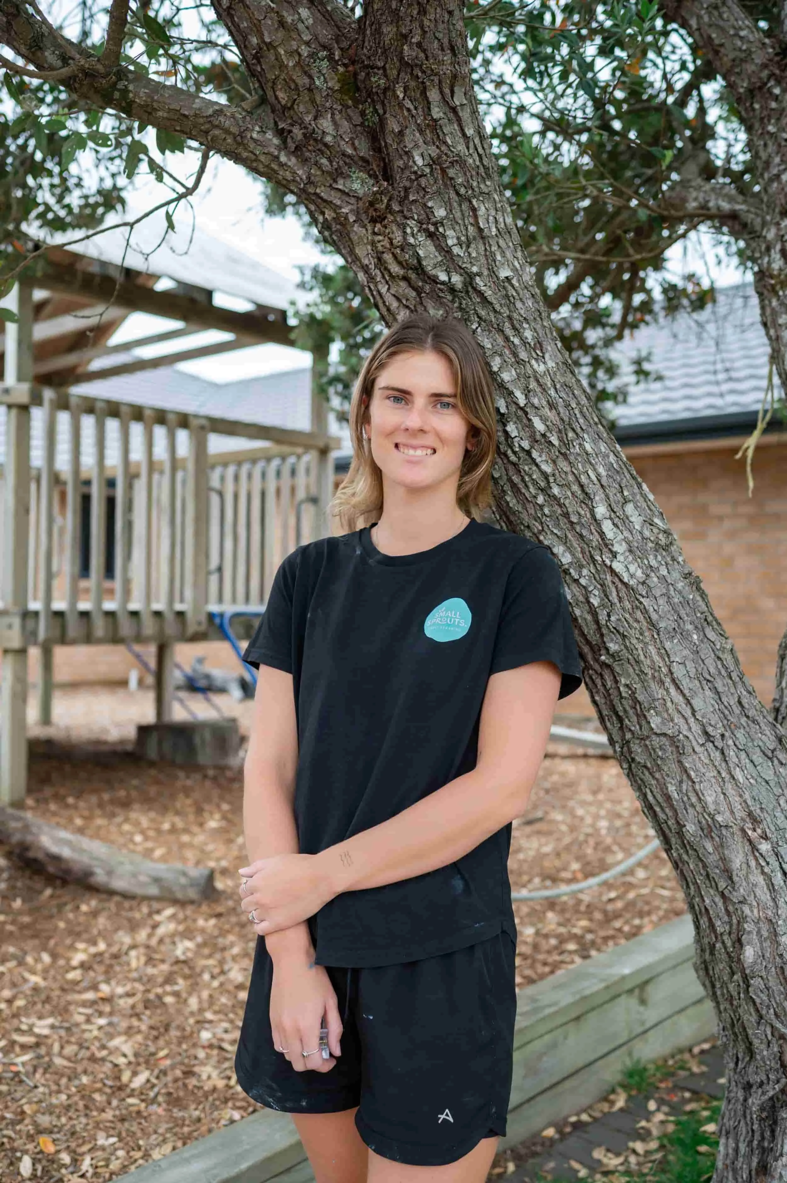 A young woman with shoulder-length hair standing outdoors against a large tree with a textured bark. She is smiling slightly and wearing a black T-shirt with a blue circular logo and black shorts, with her arms crossed. In the background, there is a wooden deck, a shed, and fallen leaves on the ground.