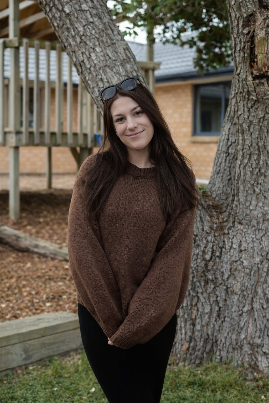 A young woman with long brown hair smiling, wearing a brown sweater and glasses on her head, standing outdoors near a large tree.