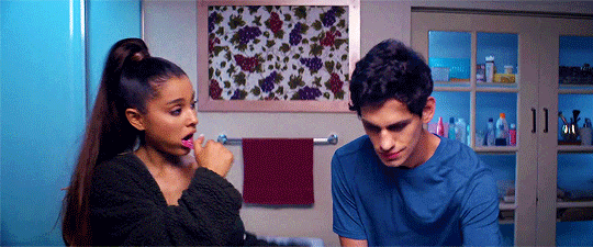 A woman and a man in a bathroom, the woman brushing her teeth and the man looking down.