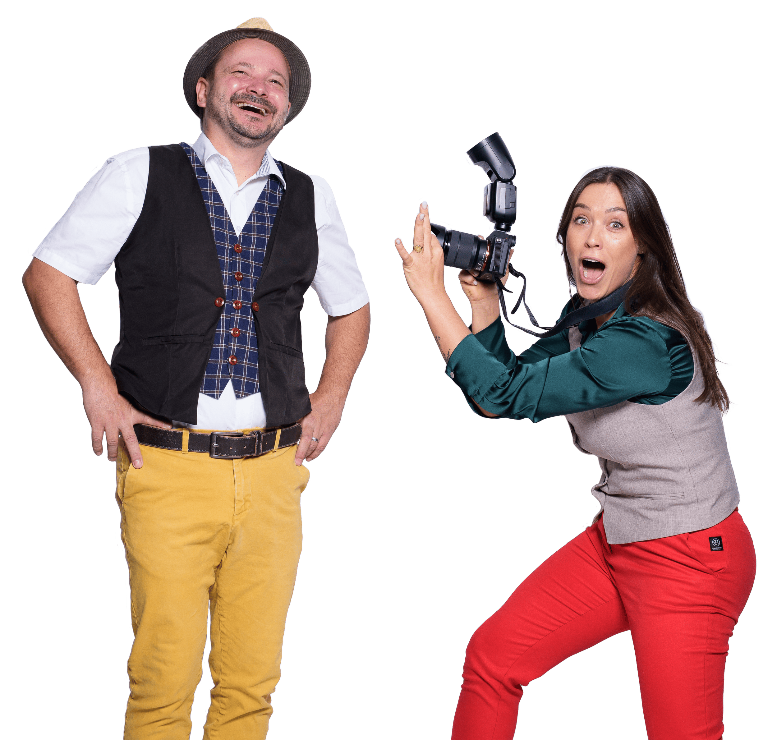 Ein lachender Mann mit Hut, Hemd, Weste und gelben Hosen steht vor einer Fotografin, die eine Kamera hält und überrascht schaut.