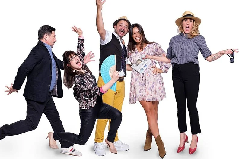 Fünf fröhliche Menschen posieren und lachen vor weißem Hintergrund, einer hält ein Handfan, eine trägt eine Sonnenbrille, und alle sind in bunter, modischer Kleidung.