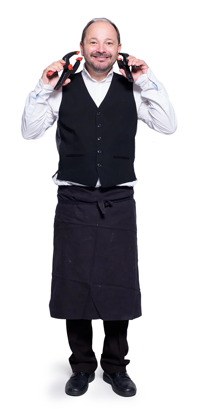 Ein humorvoller komischer Kellner mit einem langen schwarzen Schürze, weißem Hemd und schwarzen Schuhen, der zwei Schraubzwingen auf den Schultern trägt und lächelnd in die Kamera schaut.