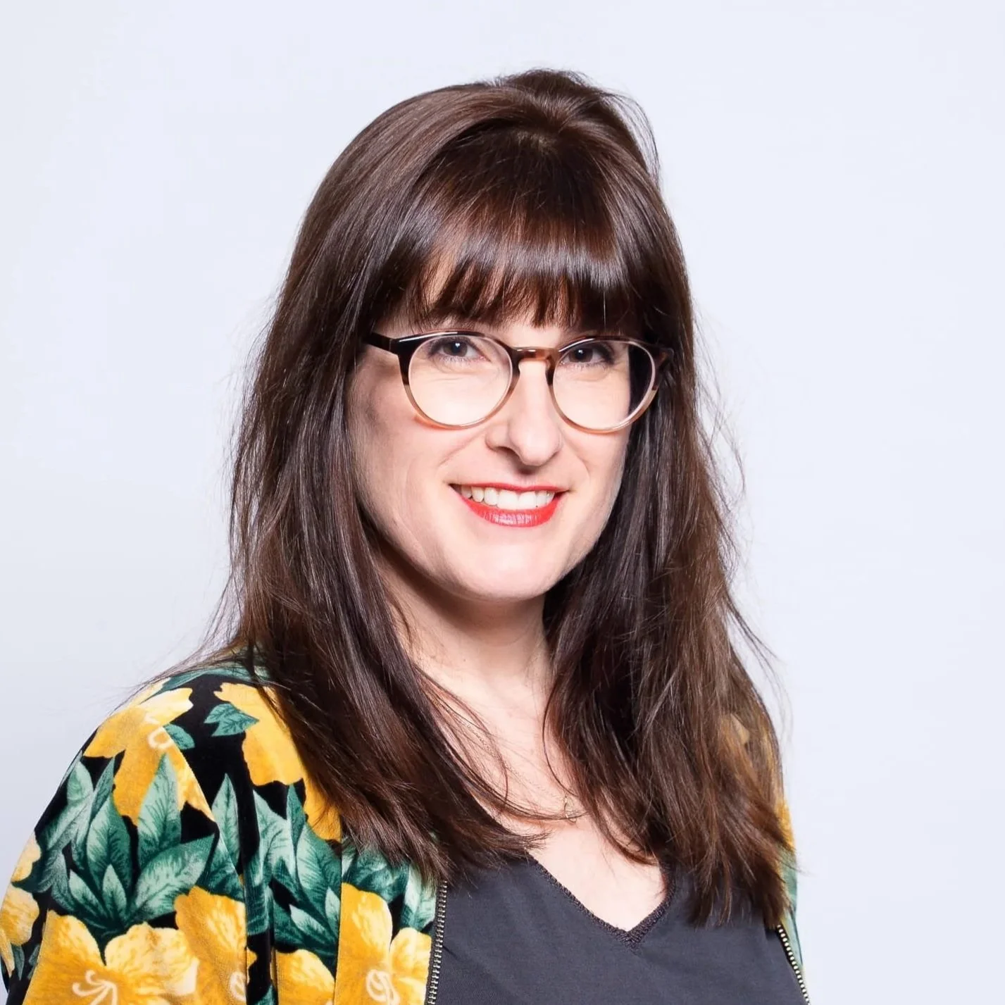 Porträt einer lächelnden Frau mit braunen Haaren, Brille und einem schwarzen Shirt, bekleidet mit einem gelben und grünen Blazer vor weißem Hintergrund.