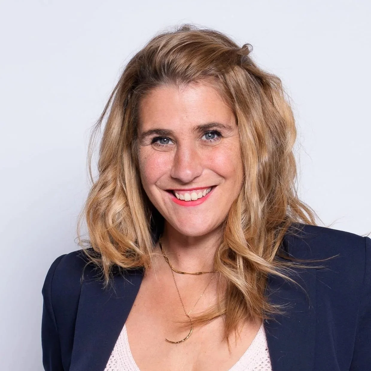 Eine lächelnde Frau mit lockigem, blondem Haar, trägt ein dunkles Jackett und eine weiße Bluse vor einem weißen Hintergrund.