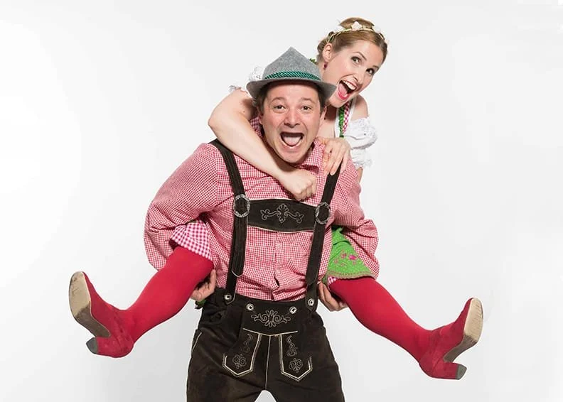 Ein Mann trägt eine Frau auf den Schultern, beide lachen fröhlich. Der Mann trägt traditionelle bayerische Kleidung, die Frau trägt ein Dirndl. Sie posieren vor einem weißen Hintergrund.
