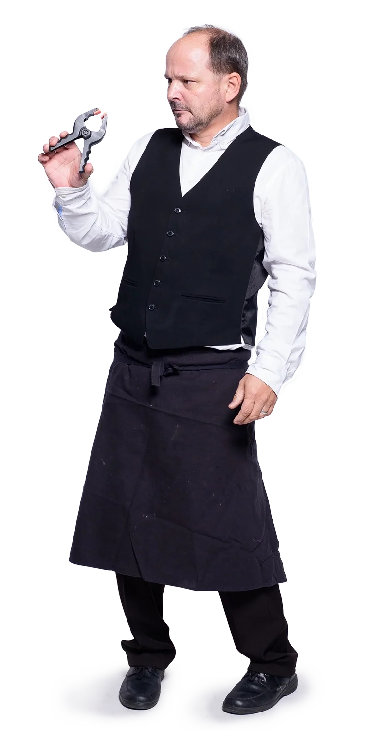 Ein Mann trägt eine weiße Hemd, eine schwarze Weste, eine schwarze Schürze und schwarze Schuhe, hält eine Klemme und schaut skeptisch, aber humorvoll.