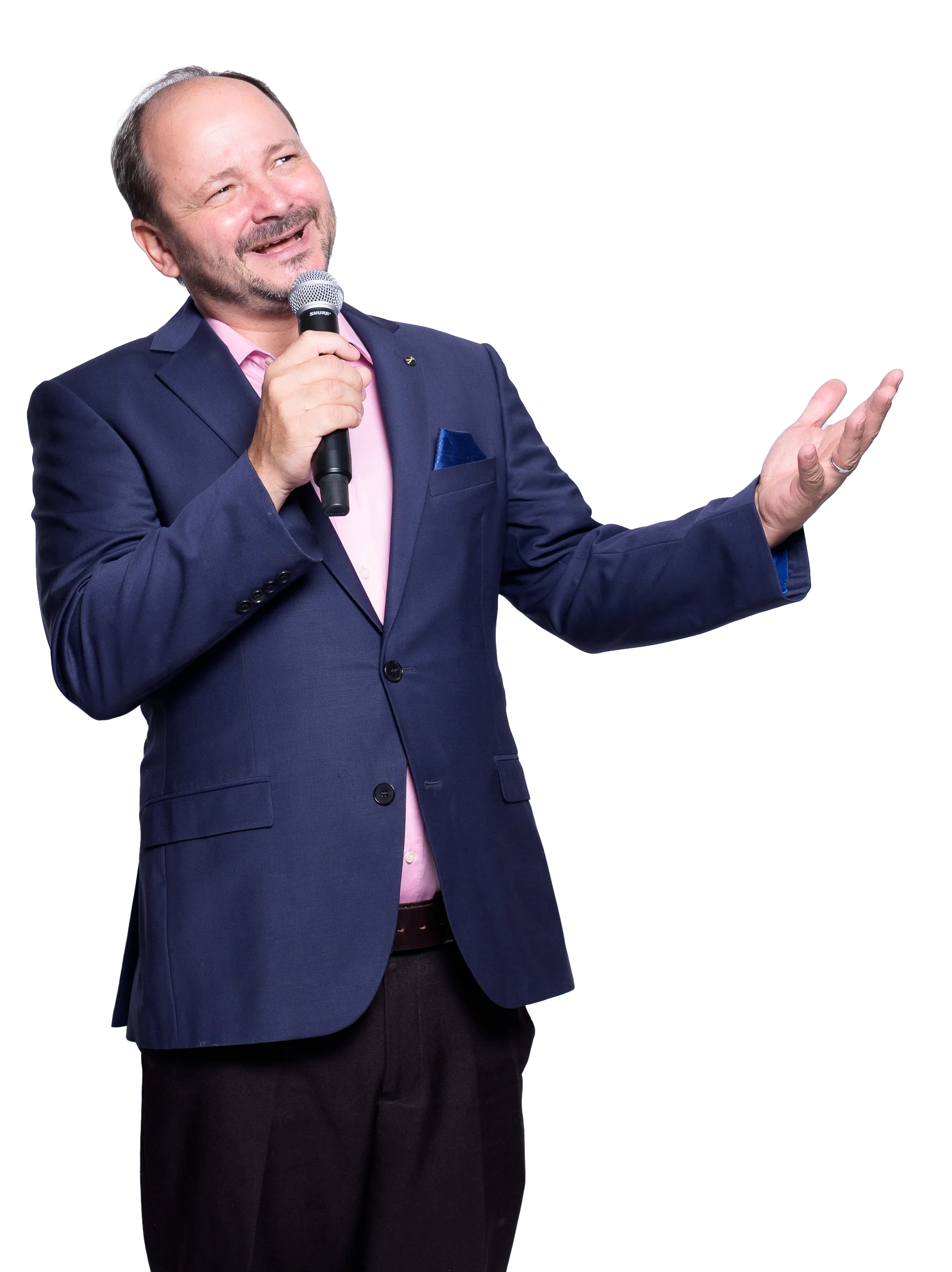 Ein komödiantischer Mann im Anzug hält ein Mikrofon und scheint eine Rede zu halten, lächelt und gestikuliert mit der anderen Hand.