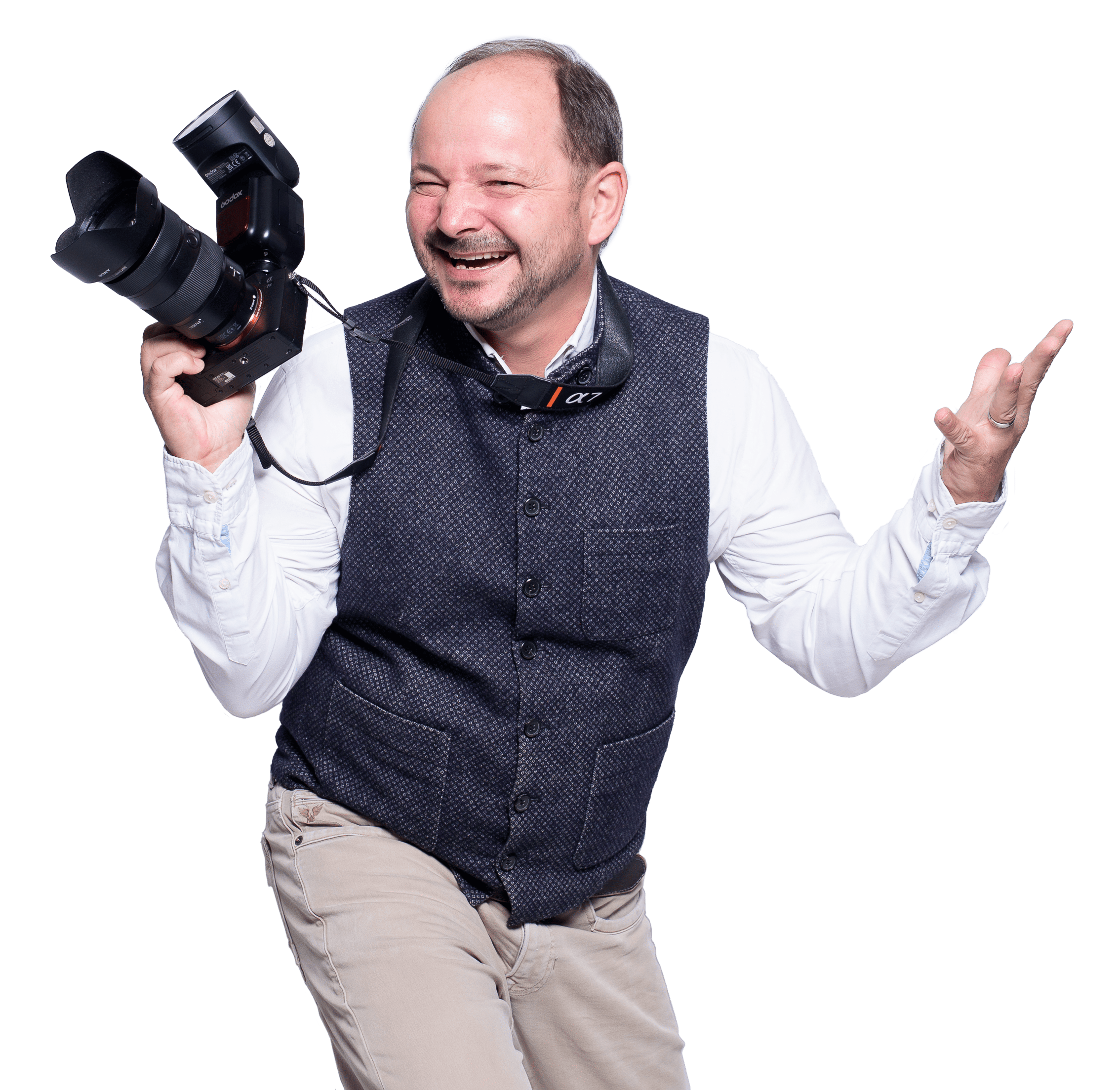 Ein lachender Mann mit Kamera in der Hand, trägt eine Brille um den Hals, Kleidung in Weiß, Dunkelblau und Beige, vor schwarzem Hintergrund.