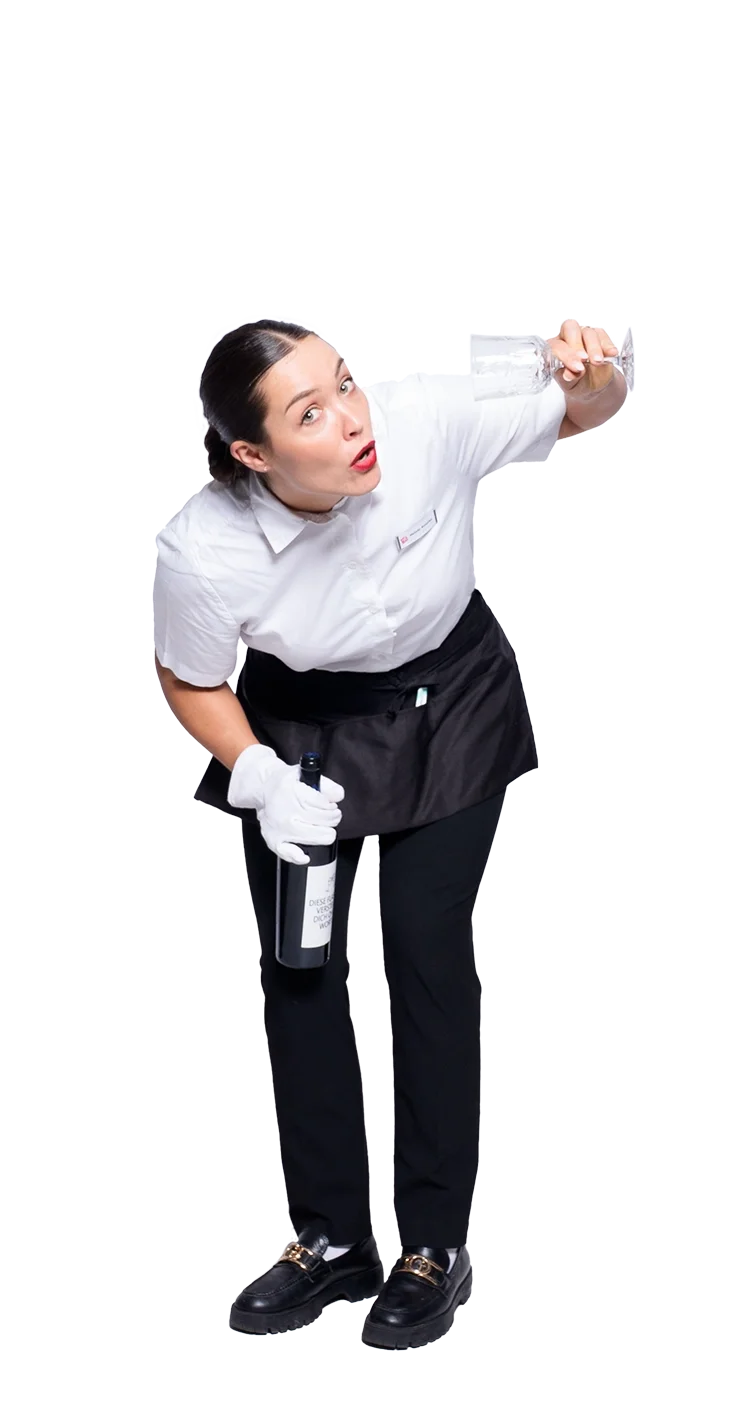 Eine weibliche humorvoller Kellnerin trägt ein weißes Hemd, schwarze Hose und Schuhe, hält eine Flasche Wein in der Hand und ein Glas Wasser, während sie neugierig in eine Richtung schaut.