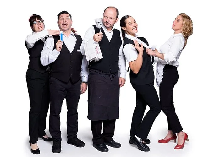 Fünf Menschen in schwarzen und weißen Kleidern, die sich gegenseitig humorvoll bedrängen.