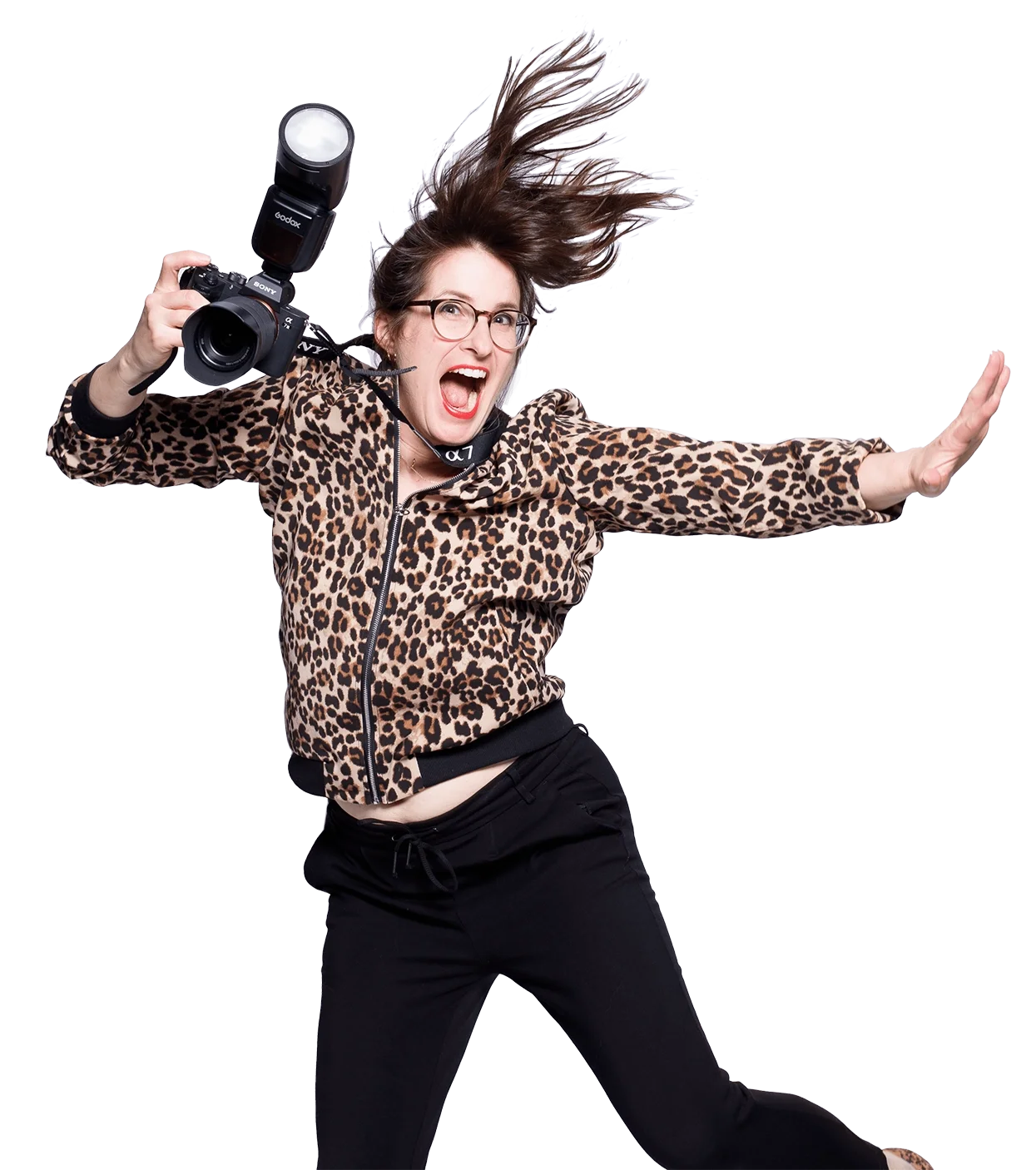 Eine Komikerin trägt eine Leo-Print Jacke, hält eine Kamera mit Blitz, hat lockiges Haar und trägt Brille. Sie posiert dynamisch mit ausgestrecktem Arm vor hellem Hintergrund.